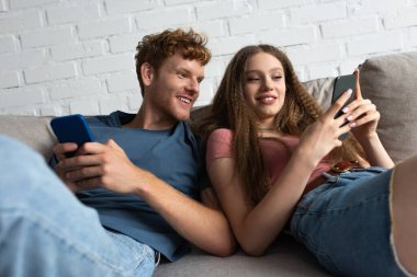 cheerful young couple using smartphones while sitting on couch in living room