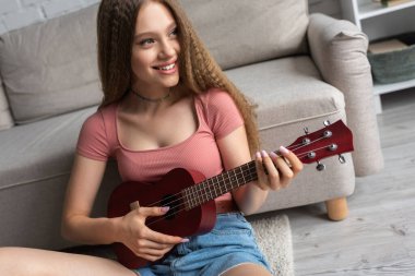 happy teenage girl in casual clothes playing ukulele in modern living room  clipart