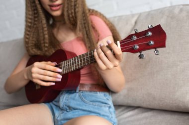 cropped view of teenage girl in casual clothes playing ukulele while sitting on couch in living room  clipart