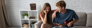 curly and redhead young man playing ukulele near happy girlfriend in living room, banner clipart