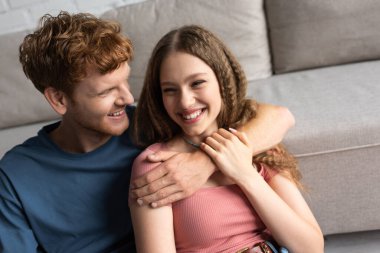 redhead and happy young man hugging and looking at cheerful girlfriend in living room 