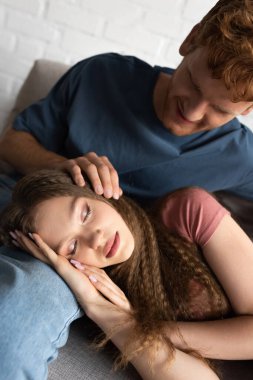 redhead and smiling young man stroking hair of sleepy girlfriend in living room 