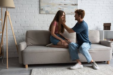 full length of redhead young man touching cheeks of happy girlfriend in living room 