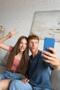 young redhead man grimacing and taking selfie with girlfriend showing peace sign in living room