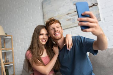 young and happy man taking selfie with positive girlfriend in living room