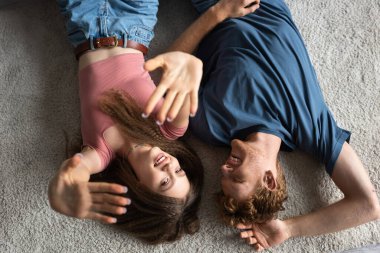 top view of happy and young man lying on carpet with cheerful girlfriend smiling while gesturing in living room 