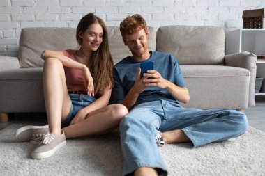 young redhead man using smartphone while sitting near cheerful girlfriend and couch in living room 