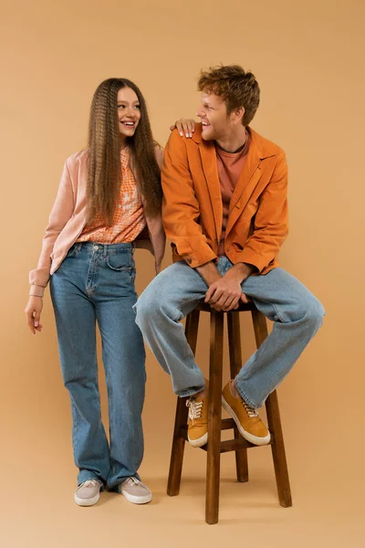 full length of young redhead man sitting on high chair and looking at girlfriend with wavy hair on beige