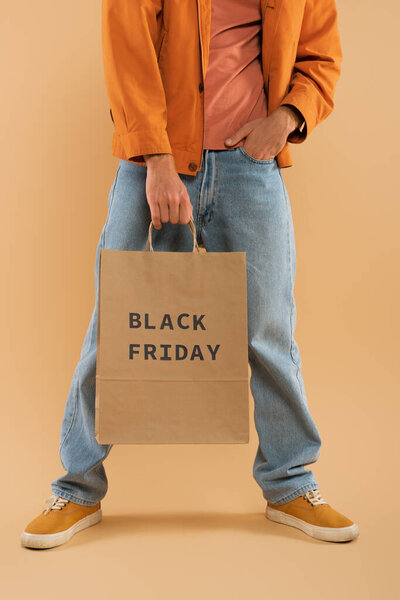 cropped view of young man holding shopping bag with black friday lettering and posing on beige