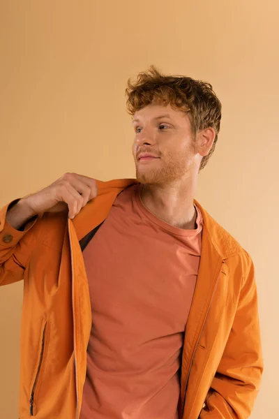 Smiling Young Man Red Hair Adjusting Orange Jacket Isolated Beige — Stok fotoğraf