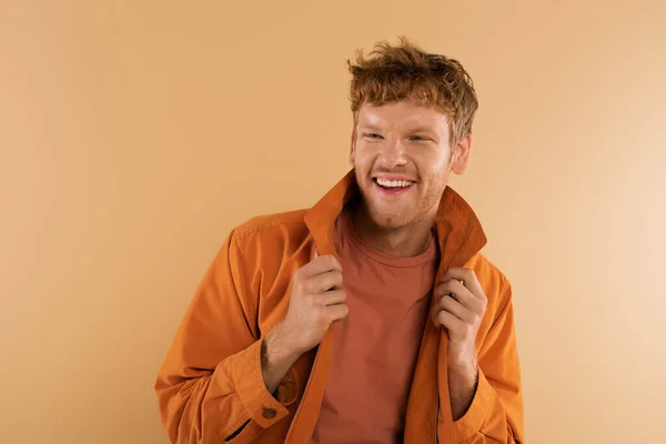 Positive Young Man Red Hair Adjusting Orange Jacket Isolated Beige —  Fotos de Stock