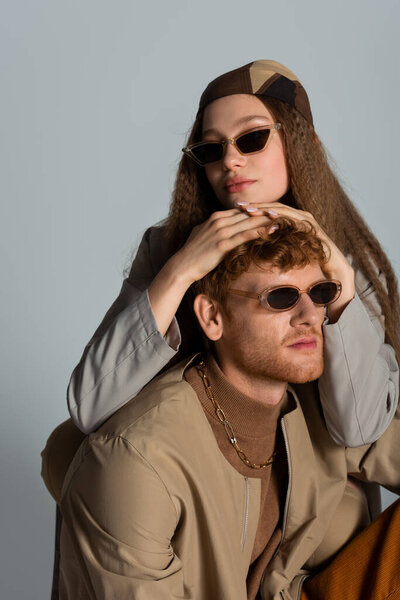young and stylish couple in sunglasses and autumnal clothes posing isolated on grey