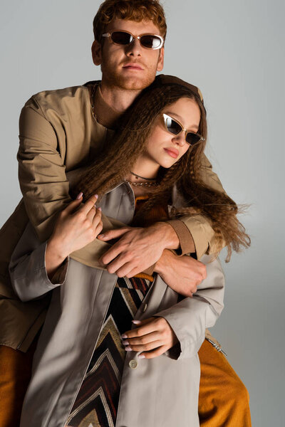 redhead young man in stylish sunglasses hugging girlfriend isolated on grey