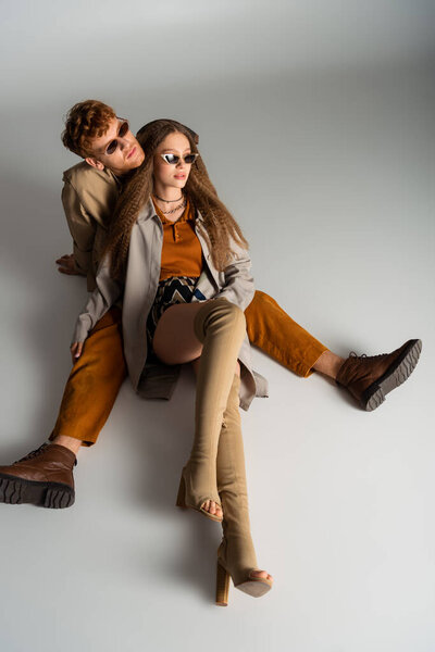 young stylish couple in sunglasses and trendy outfits sitting on grey