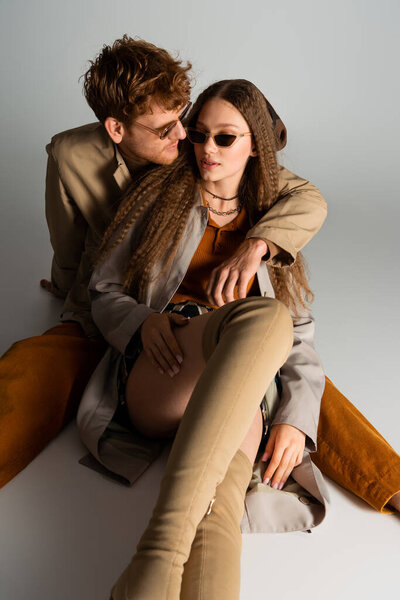 stylish young man in sunglasses hugging girlfriend and sitting on grey