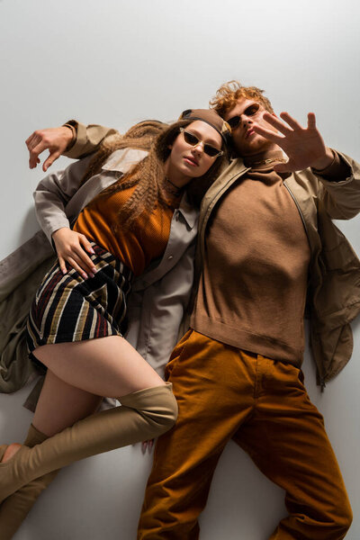 top view of stylish young couple in autumnal outfits and sunglasses lying together isolated on grey 