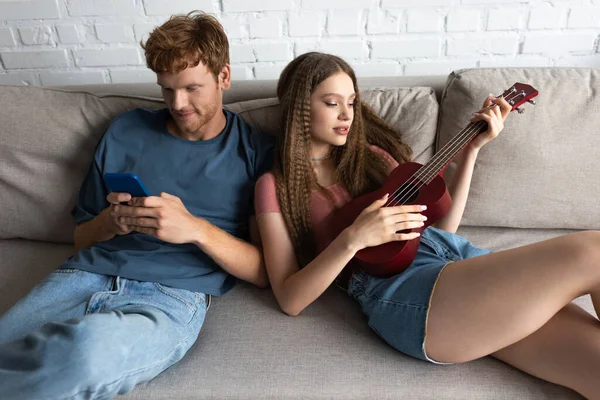 Happy Redhead Man Using Smartphone While Girlfriend Playing Ukulele Couch —  Fotos de Stock