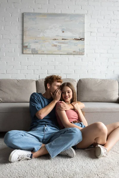 redhead and happy young man hugging and whispering in ear of cheerful girlfriend in modern living room 