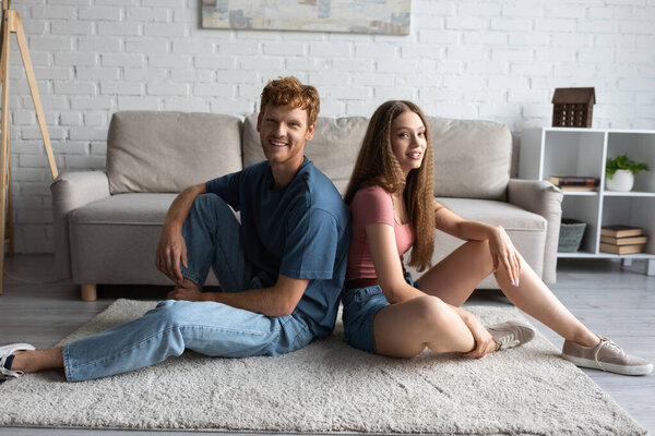 full length of happy young couple sitting on carpet and looking at camera in living room 
