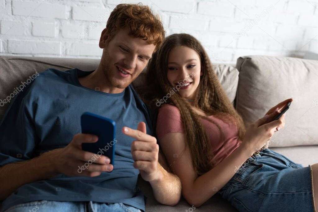 young man pointing at mobile phone while sitting on couch with happy girlfriend in living room