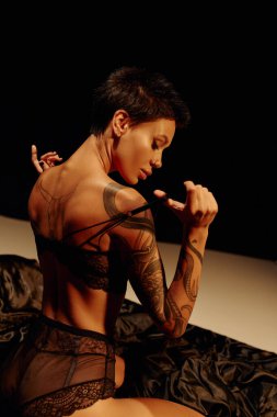 tutkulu dövmeli kadın siyah arka planda ipek yatakta otururken sütyenini çıkarıyor.
