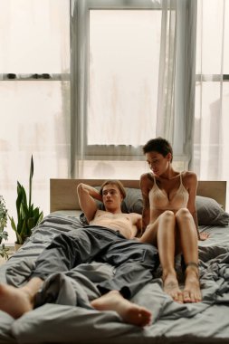 Pijamalı gömleksiz bir adam yatakta seksi bir kız arkadaşın yanında yatıyor. 