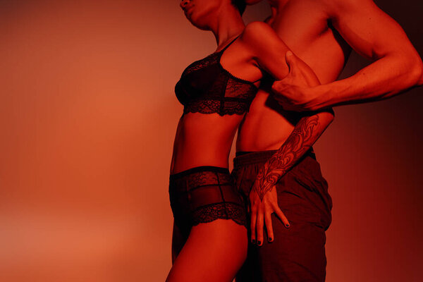 partial view of sexy woman in black lace lingerie near shirtless man on red background