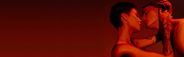 Вид сбоку молодой татуированной женщины, целующейся и обнимающей дерзкого мужчину, изолированного на красном баннере