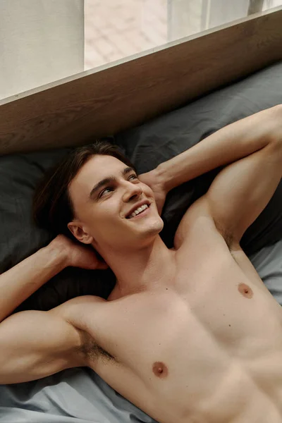 top view of sexy young man looking away and smiling while lying on bed with hands behind head