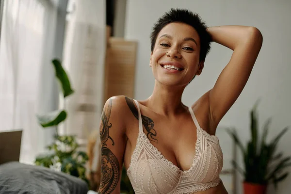 happy tattooed woman in beige bra holding hand behind head and smiling at camera