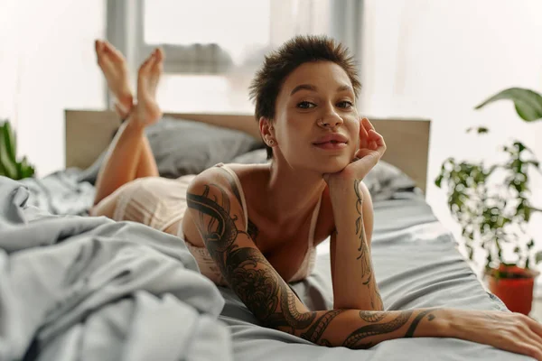 Tattooed woman in lingerie looking at camera while lying on grey bedding at home