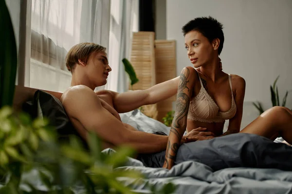 Young man in pajama pants touching tattooed girlfriend in bra on bed