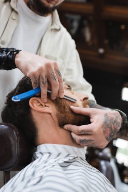 tattooed hairdresser shaving cheek of client with straight razor clipart