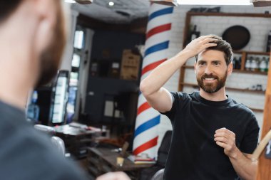 Esmer sakallı adam aynaya bakarken gülümsüyor ve ön plandaki bulanık saçlara dokunuyor.