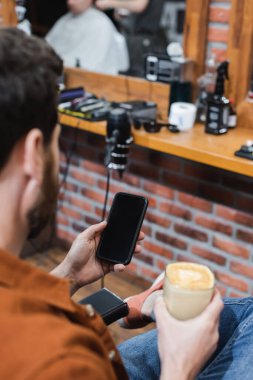 Berber dükkanında boş ekranlı akıllı telefon tutan bir bardak cappuccino ile bulanık müşteri görüntüsü
