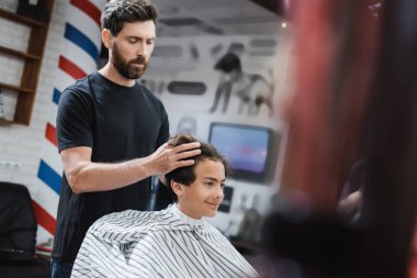 Bearded hairstylist styling hair of smiling teenage boy in beauty salon  clipart