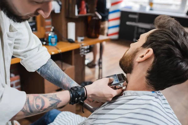 Wytatuowany Fryzjer Skórzanej Bransoletce Golący Szyję Brodatego Klienta Brzytwą Elektryczną — Zdjęcie stockowe