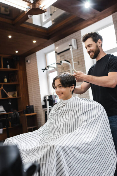 bearded barber working with teenage client sitting in hairdressing cape in beauty salon