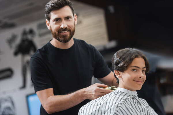 Smiling hairdresser holding trimmer near client and looking at camera in beauty salon 