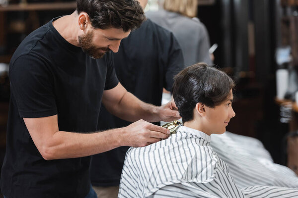 Bearded barber holding hair clipper near neck of teenager in beauty salon 