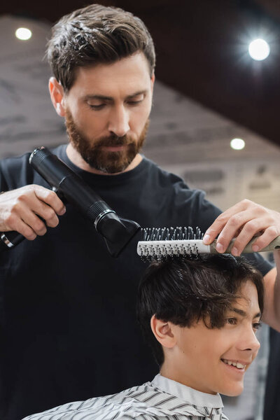 Barber drying and combing hair of smiling teen boy in beauty salon 