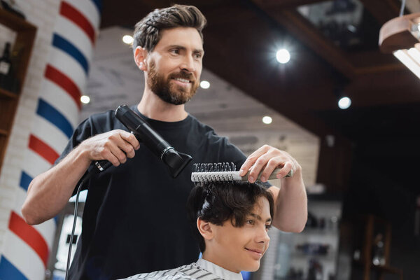 Positive hairstylist combing and drying hair of teenager in beauty salon  