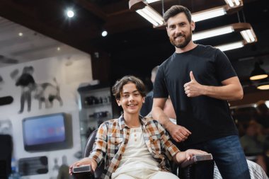Gülümseyen genç çocuk kuaförün yanındaki kameraya bakıyor. Güzellik salonundaki kameraya başparmağını gösteriyor. 