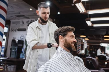 Berber dükkanında berberin yanında oturan kuaför pelerinli neşeli adam. 