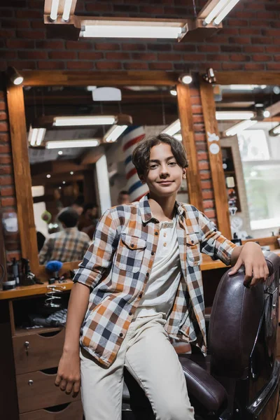 ポジティブなティーン男の子見ますカメラ近くArmchairでぼやけたBarbershop — ストック写真