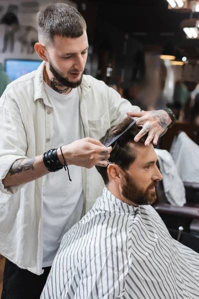 Tattooed barber combing hair of bearded customer in barbershop