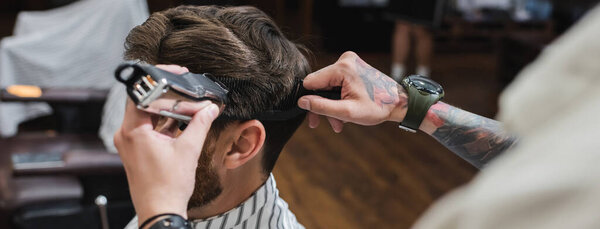 Tattooed barber combing and trimming hair of man in barbershop, banner 