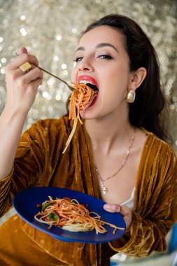 Kadife elbiseli kadın lezzetli spagetti yiyor ve parlak gümüş arka planda kameraya bakıyor.
