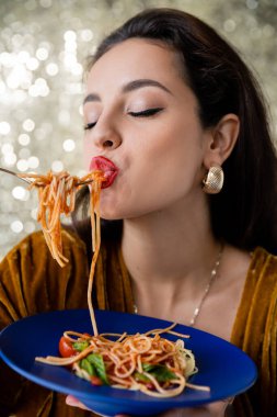 Makyajlı ve gözleri kapalı esmer bir kadın ışıltılı arka planda lezzetli spagetti yiyor.