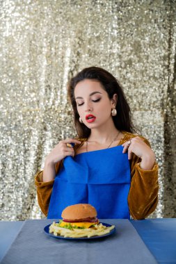 Baştan çıkarıcı kadın tabağın yanında mavi peçeteyle patates kızartması ve parlak gümüş arka planda hamburger tutuyor.